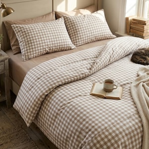 May include: A bedding set featuring a light brown and white gingham check pattern on the duvet cover and two matching pillowcases. An open book and a ceramic mug sit on the bed, alongside a folded cream-coloured knit throw blanket.