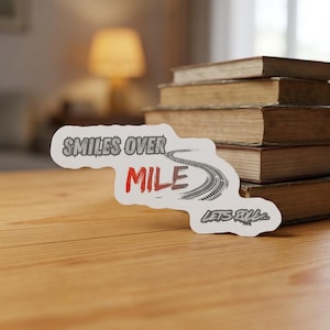 Könnte beinhalten: Ein weißer, konturgestanzter Aufkleber mit dem Text SMILES OVER MILE in grauer und roter Schrift, ergänzt durch eine schwarze Reifenspur-Grafik und den kleineren Text LETS ROLL in einer stilisierten grauen Schrift auf weißem Grund.