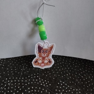 May include: A small, brown and white cartoon animal charm with green and yellow beads on a white string. The charm is attached to a white string with a knot at the top.