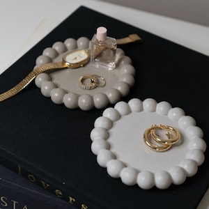 May include: Two gray concrete ring dishes with raised dots around the edges. One dish has a gold watch and two gold rings, the other has a pair of gold hoop earrings. The dishes are on top of two black books.