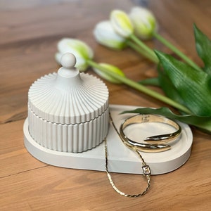 May include: A white ceramic trinket dish with a fluted design, featuring a lid with a dome shape. The dish is on a gray oval tray with a gold chain necklace and a gold bangle bracelet.