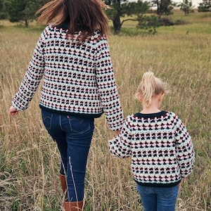 Könnte beinhalten: Eine Frau und ein Kind gehen durch ein Feld, beide tragen passende Jacken mit Muster. Die Jacken haben ein geometrisches Design in Schwarz, Weiß und Rot. Sie tragen auch blaue Jeans und braune Stiefel. Die Szene spielt in einem grasbewachsenen Feld mit Bäumen im Hintergrund.