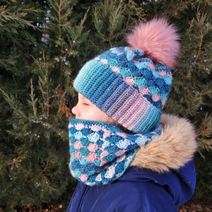 May include: A blue, pink, and purple crocheted hat and cowl set. The hat has a large pink pom-pom. The cowl is wrapped around the neck. The set is designed for warmth in cold weather. The set is a cozy winter accessory.