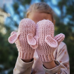 May include: A pair of pink knitted mittens with ear-like flaps. The mittens are made with a textured stitch and are perfect for keeping hands warm in cold weather.