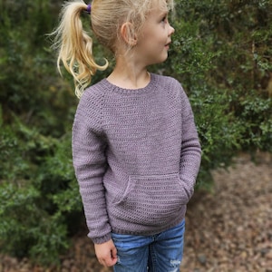 May include: A lavender-colored, crocheted sweater with a front pocket. The long-sleeved pullover has a ribbed hem and cuffs. The sweater is paired with blue jeans. The child is wearing the sweater.