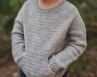 May include: A light gray, crocheted sweater with a round neckline and long sleeves. The sweater features a front pocket and a ribbed hem and cuffs. The yarn has a textured appearance. The sweater is designed for children.