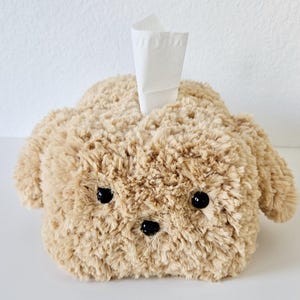 May include: A brown plush tissue box cover shaped like a dog with black eyes and a black nose. A white tissue is sticking out of the top.
