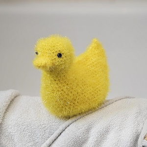 May include: A yellow knitted duck toy with black eyes. The duck is sitting on a white towel.