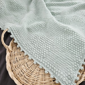 May include: A soft, sage green crocheted blanket with a textured stitch pattern and a scalloped edge. The blanket is folded and displayed on a woven basket.