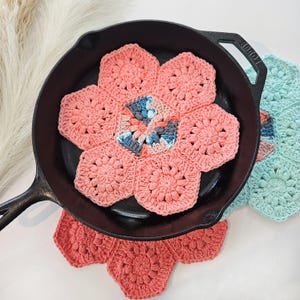 Pode incluir: Uma frigideira de ferro fundido preta contém um descanso de panela em forma de flor de crochê na cor coral. O descanso de panela é feito de peças hexagonais, com um centro azul e cinza. Outro descanso de panela coral e um turquesa também são visíveis.