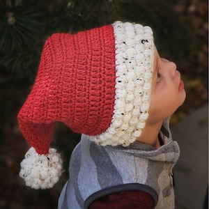 May include: A red and white crocheted Santa hat with a white pom-pom on the end. The hat is worn by a child.