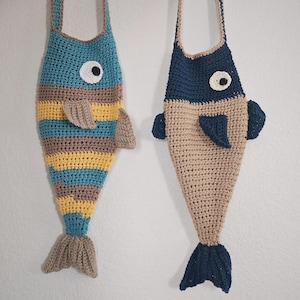 Op de afbeelding: Twee gehaakte tassen in de vorm van een vis die naast elkaar hangen. De linker tas heeft horizontale strepen in teal, geel en beige met een wit en zwart oog. De rechter tas is beige met een donkerblauw bovendeel, vinnen en staart met een oog.