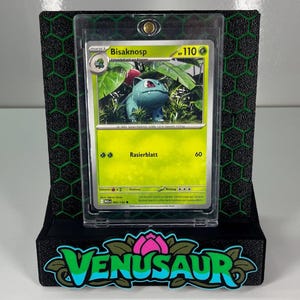 May include: A Bisaknosp trading card encased in a clear protective magnetic holder, mounted on a black display stand with a green hexagonal pattern and the word VENUSAUR in large green and blue lettering at the base.