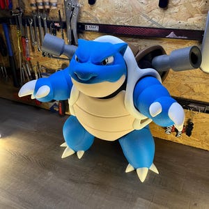 May include: A blue and white Blastoise figure, a popular character from the Pok&eacute;mon franchise. The figure has a light tan shell, blue body, and two gray cannons on its back. The figure is standing on a wooden surface.