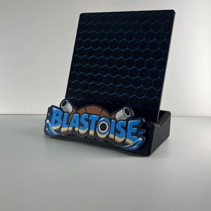 Puede incluir: Un soporte para tel&eacute;fono negro con un panel trasero vertical con patr&oacute;n de panal azul. La base frontal muestra la palabra BLASTOISE en letras azules y amarillas, acentuada por dos ca&ntilde;ones cil&iacute;ndricos grises y un dise&ntilde;o de caparaz&oacute;n marr&oacute;n.