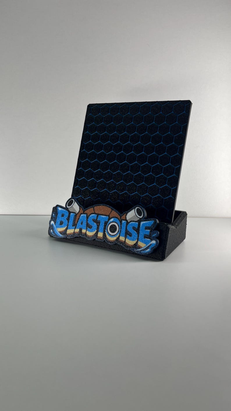Puede incluir: Un soporte para tel&eacute;fono negro con un panel trasero vertical con patr&oacute;n de panal azul. La base frontal muestra la palabra BLASTOISE en letras azules y amarillas, acentuada por dos ca&ntilde;ones cil&iacute;ndricos grises y un dise&ntilde;o de caparaz&oacute;n marr&oacute;n.