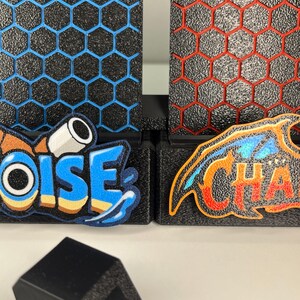 Puede incluir: Dos paneles negros texturizados con un patr&oacute;n de rejilla hexagonal, uno con contornos azules y otro con contornos rojos. Los paneles muestran parches bordados coloridos con el texto OISE y CHA. Un peque&ntilde;o clip de pl&aacute;stico negro en forma de U se sit&uacute;a delante.
