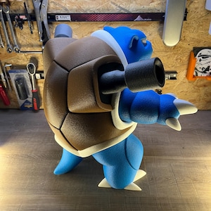 May include: A blue and brown Blastoise figure, a popular character from the Pok&eacute;mon franchise. The figure has a brown shell, two black cannons, and white accents. The figure is standing on a wooden surface.