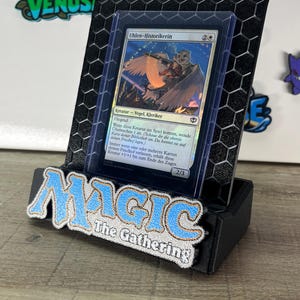 Op de afbeelding: Een zwarte 3D-geprinte displaystandaard met een zeshoekig patroon waarin een Magic: The Gathering-ruilkaart met de titel Uhlen-Historikerin staat. De standaard heeft het blauw-witte Magic: The Gathering-logo op de voorzijde.