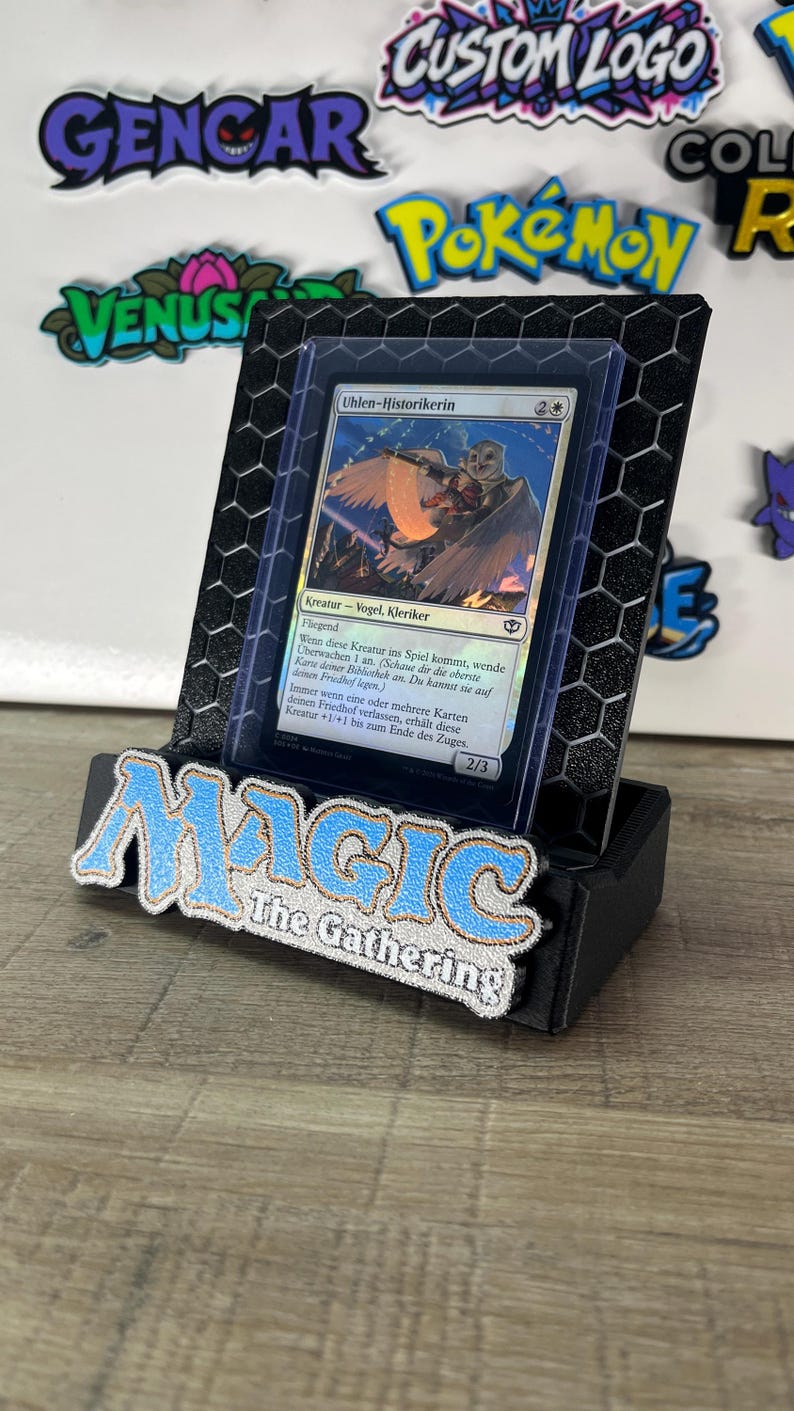 Op de afbeelding: Een zwarte 3D-geprinte displaystandaard met een zeshoekig patroon waarin een Magic: The Gathering-ruilkaart met de titel Uhlen-Historikerin staat. De standaard heeft het blauw-witte Magic: The Gathering-logo op de voorzijde.