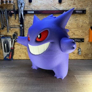 May include: A 3D-printed purple Gengar figure with a wide grin, red eyes, and spiky features. The figure is standing on a wooden surface. The figure is a popular character from the Pokemon franchise.