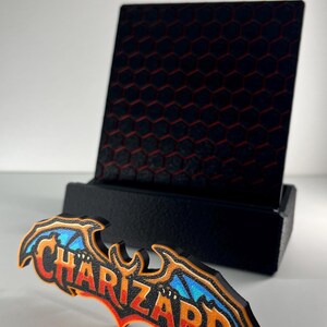 May include: A black display stand with a hexagonal pattern holds a decorative sign featuring the word CHARIZARD in orange and red lettering with blue wing accents, all outlined in black, positioned on a plain white surface.
