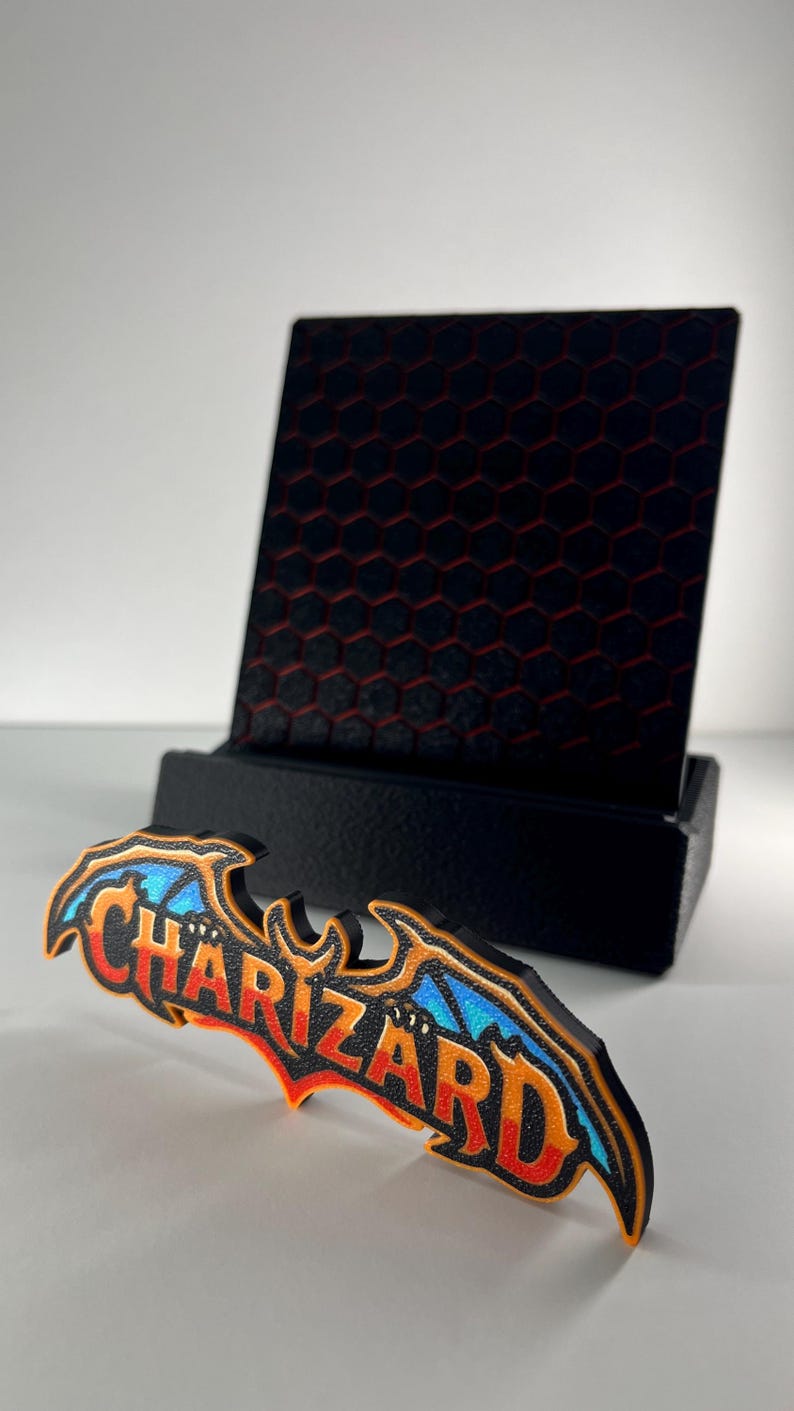May include: A black display stand with a hexagonal pattern holds a decorative sign featuring the word CHARIZARD in orange and red lettering with blue wing accents, all outlined in black, positioned on a plain white surface.
