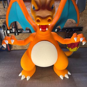 May include: A large, orange Charizard figure with blue wings and white claws. The figure has a white belly and a detailed face with blue eyes and an open mouth. The figure is posed with its arms outstretched.