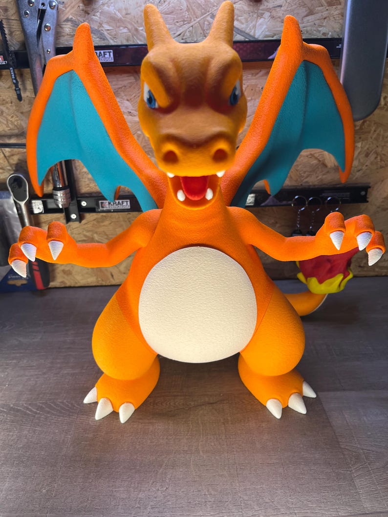 May include: A large, orange Charizard figure with blue wings and white claws. The figure has a white belly and a detailed face with blue eyes and an open mouth. The figure is posed with its arms outstretched.
