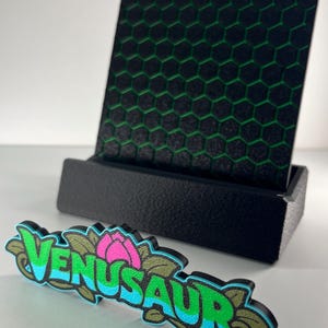 May include: A black display stand holding a square plaque with a green honeycomb pattern. In front sits a decorative sign featuring the word VENUSAUR in bright blue and green letters with a pink flower icon and leaf accents.