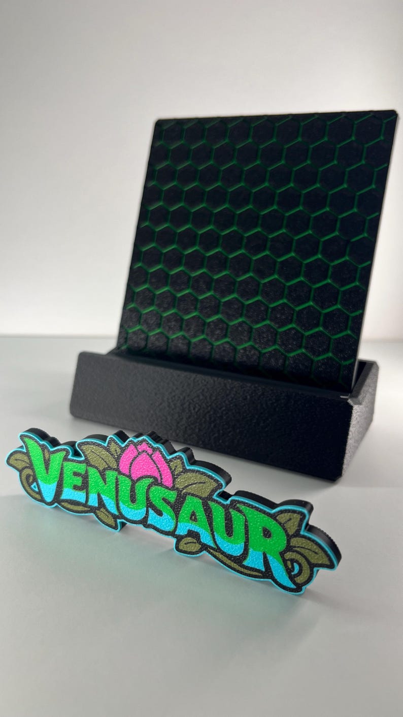 May include: A black display stand holding a square plaque with a green honeycomb pattern. In front sits a decorative sign featuring the word VENUSAUR in bright blue and green letters with a pink flower icon and leaf accents.