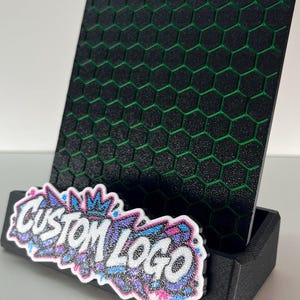 K&ouml;nnte beinhalten: Ein schwarzer Pr&auml;sentationsst&auml;nder mit texturiertem Wabenmuster und gr&uuml;nen Akzenten. Auf der Vorderseite befindet sich eine wei&szlig;-blau-rosa glitzernde Grafikplatte mit dem Text CUSTOM LOGO in einer Schriftart im Graffiti-Stil.