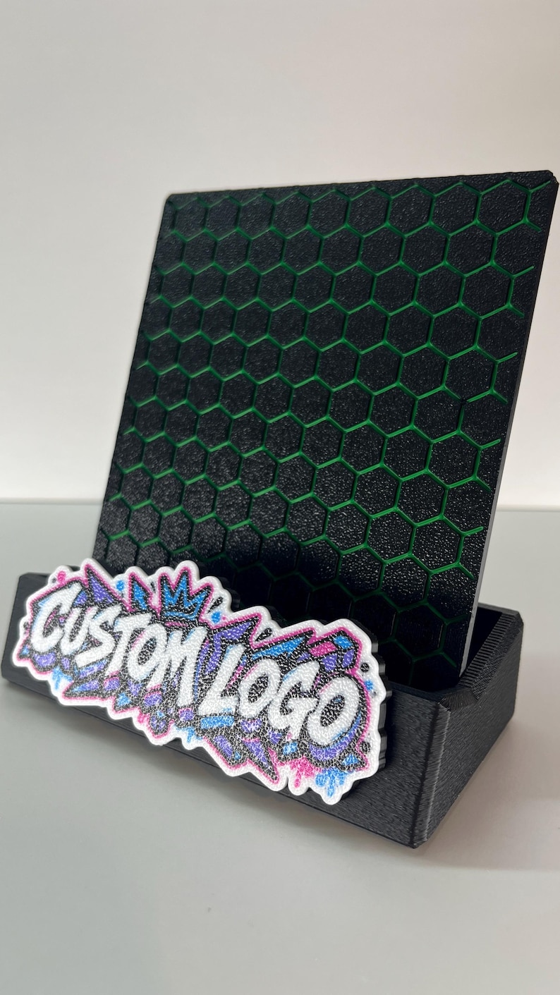 K&ouml;nnte beinhalten: Ein schwarzer Pr&auml;sentationsst&auml;nder mit texturiertem Wabenmuster und gr&uuml;nen Akzenten. Auf der Vorderseite befindet sich eine wei&szlig;-blau-rosa glitzernde Grafikplatte mit dem Text CUSTOM LOGO in einer Schriftart im Graffiti-Stil.