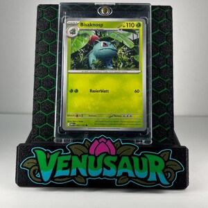 May include: A Bisaknosp trading card in a clear protective case, displayed on a black stand with a green honeycomb pattern and the word VENUSAUR in stylized blue and green lettering with a pink flower icon above the text.