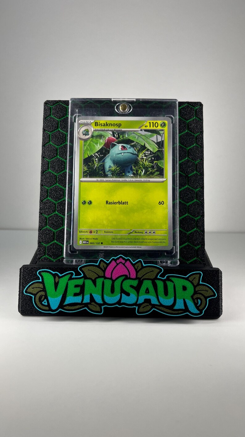 May include: A Bisaknosp trading card in a clear protective case, displayed on a black stand with a green honeycomb pattern and the word VENUSAUR in stylized blue and green lettering with a pink flower icon above the text.