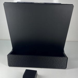 Op de afbeelding: Een zwarte 3D-geprinte displaystandaard met een breed, plat achterpaneel en een rechthoekige basis met een getextureerd oppervlak. Een apart, kleiner zwart rechthoekig blok met een inkeping in het midden staat voor de standaard.