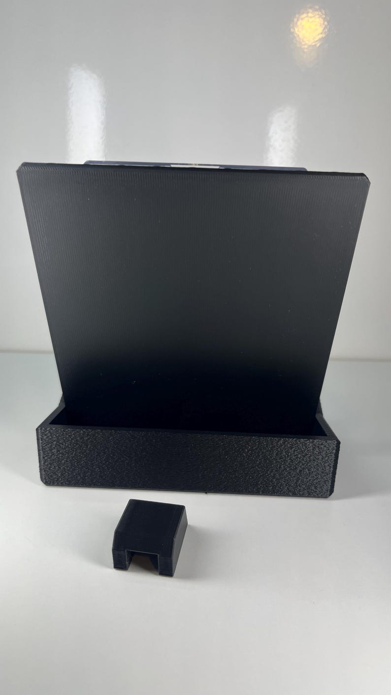 Op de afbeelding: Een zwarte 3D-geprinte displaystandaard met een breed, plat achterpaneel en een rechthoekige basis met een getextureerd oppervlak. Een apart, kleiner zwart rechthoekig blok met een inkeping in het midden staat voor de standaard.