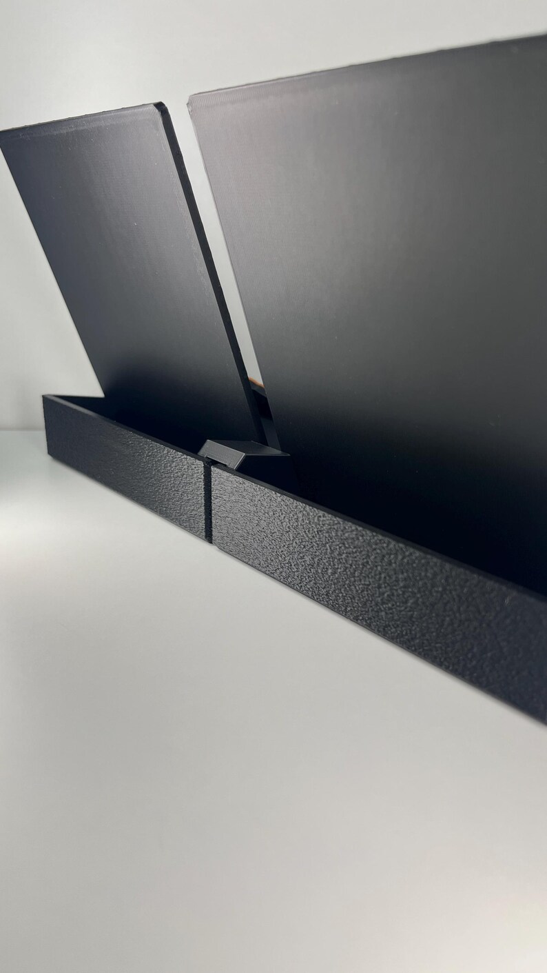 Puede incluir: Un soporte de exhibici&oacute;n negro con dos paneles rectangulares verticales de superficie lisa, sujetos por una base texturizada de perfil bajo. La estructura est&aacute; compuesta de material negro mate, ofreciendo un soporte estable.