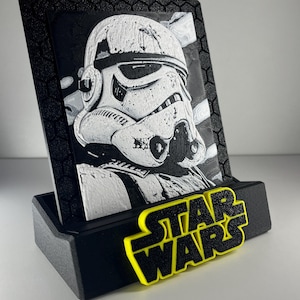K&ouml;nnte beinhalten: Eine schwarz-wei&szlig;e 3D-gedruckte Plakette mit einem Stormtrooper-Helm-Design, eingefasst in einen schwarzen Rahmen mit Sechseckmuster. Die Plakette steht auf einem schwarzen Sockel, der vorne das gelb-schwarze Star Wars-Logo zeigt.