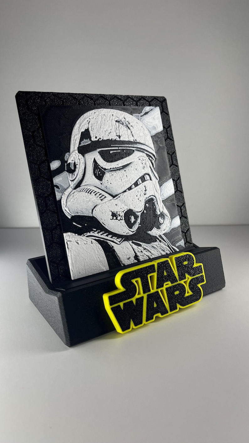 K&ouml;nnte beinhalten: Eine schwarz-wei&szlig;e 3D-gedruckte Plakette mit einem Stormtrooper-Helm-Design, eingefasst in einen schwarzen Rahmen mit Sechseckmuster. Die Plakette steht auf einem schwarzen Sockel, der vorne das gelb-schwarze Star Wars-Logo zeigt.