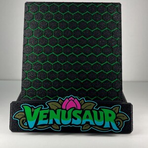 May include: A black display stand featuring a textured honeycomb pattern with green accents. The front base displays the text VENUSAUR in blue letters, accented by a pink flower graphic and green leaves, all set against a solid black background.