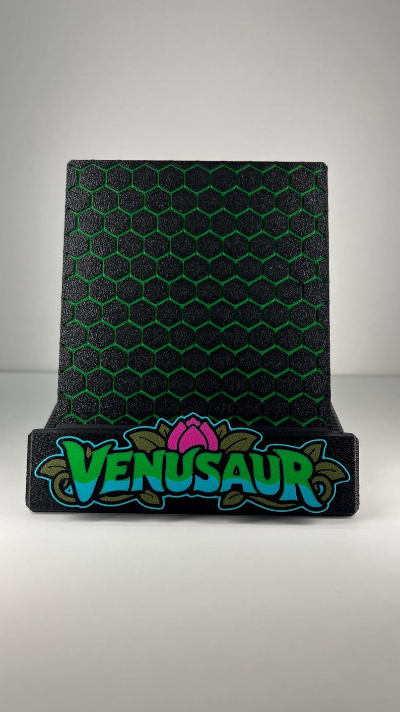 May include: A black display stand featuring a textured honeycomb pattern with green accents. The front base displays the text VENUSAUR in blue letters, accented by a pink flower graphic and green leaves, all set against a solid black background.
