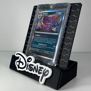 May include: A black 3D-printed display stand featuring a hexagonal pattern and the white Disney logo on the front. The stand holds a Gengar Pok&eacute;mon trading card encased in a clear protective plastic sleeve, showcasing the character artwork.