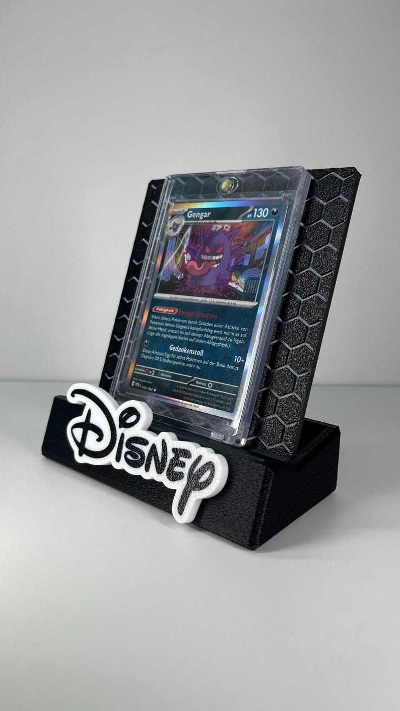 May include: A black 3D-printed display stand featuring a hexagonal pattern and the white Disney logo on the front. The stand holds a Gengar Pok&eacute;mon trading card encased in a clear protective plastic sleeve, showcasing the character artwork.