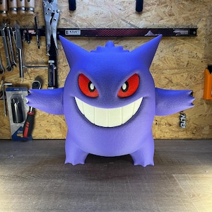 May include: A large, purple, three-dimensional figure of a cartoon character with a wide grin, red eyes, and small horns. The figure has outstretched arms and is standing on a wooden surface. The background includes tools and a wooden wall.