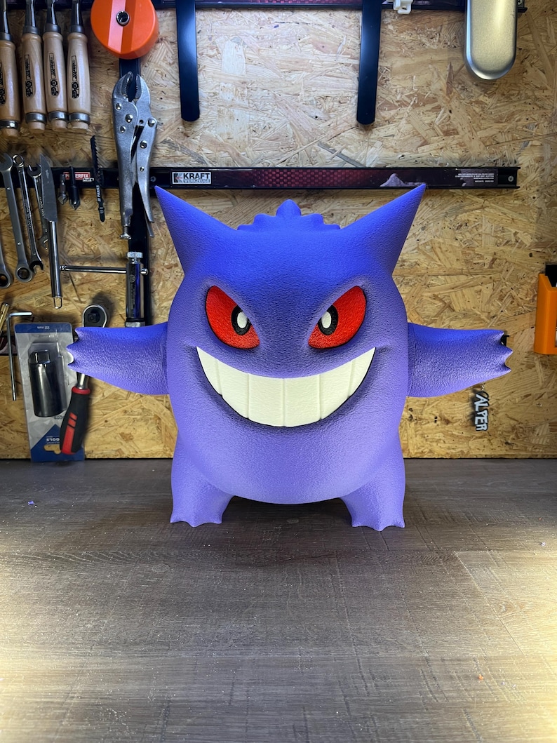 May include: A large, purple, three-dimensional figure of a cartoon character with a wide grin, red eyes, and small horns. The figure has outstretched arms and is standing on a wooden surface. The background includes tools and a wooden wall.