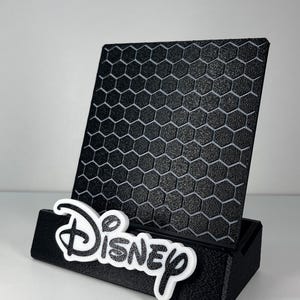 May include: A black phone stand featuring a textured honeycomb pattern on the back panel. The base of the stand is black and displays the word Disney in white, raised, stylized lettering attached to the front edge of the holder.
