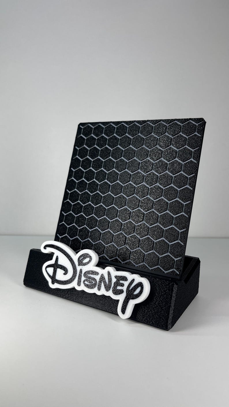 May include: A black phone stand featuring a textured honeycomb pattern on the back panel. The base of the stand is black and displays the word Disney in white, raised, stylized lettering attached to the front edge of the holder.