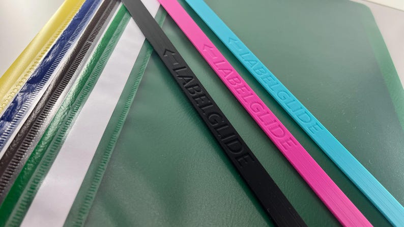 Peut inclure: Une collection de bandes en plastique fines, plates et rectangulaires de diff&eacute;rentes couleurs, dont jaune, bleu, noir, blanc, vert, rose et cyan. Les bandes noire, rose et cyan portent le texte en relief LABELGLIDE avec une ic&ocirc;ne de fl&egrave;che.