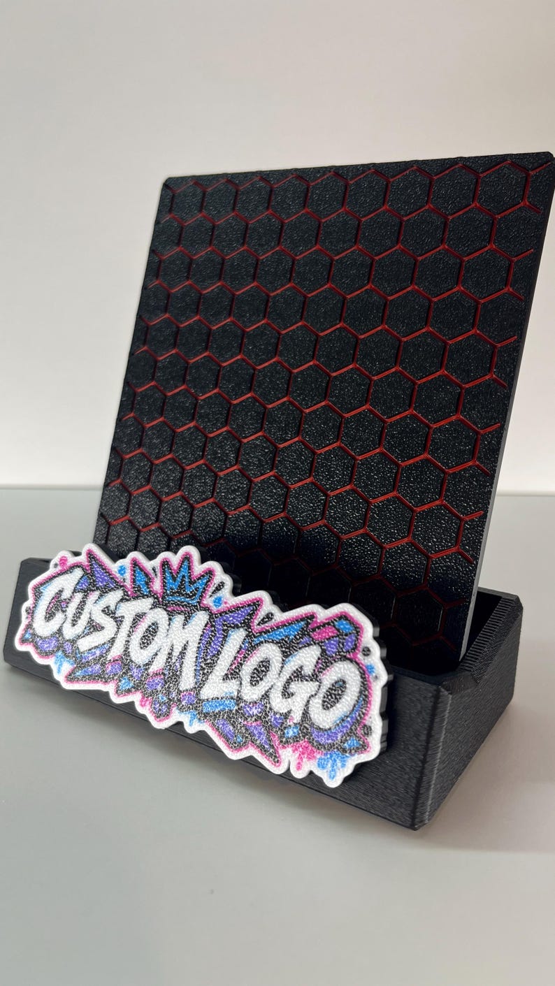 K&ouml;nnte beinhalten: Ein schwarzer Handyhalter mit texturiertem Sechseckmuster und roten Akzenten in den Vertiefungen. Die Vorderseite zeigt einen glitzernden Aufn&auml;her in Wei&szlig;, Blau und Pink mit dem Text CUSTOM LOGO in einer markanten Schriftart.